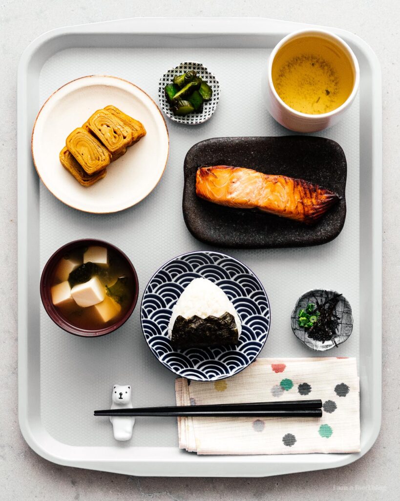 An overhead photo of a typical Japanese breakfast plate: including micro-servings of fish, miso soup, pickled vegetables, rice, and sweet, rolled omelets
