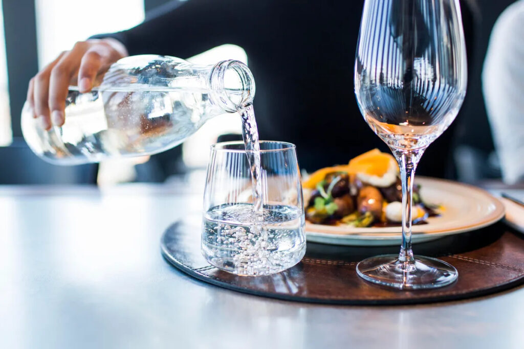 Person pouring a glass of water with their meal, next to an empty wine glass